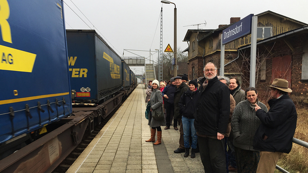 Ein Güterzug fährt durch den Bahnhalt Dahnsdorf, während eine Gruppe interessierter Personen auf dem Bahnsteig diesem zuschaut, Foto: Michael Jungclaus MdL